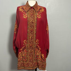 May include: A long-sleeved, red button-down shirt with an ornate gold and brown floral pattern. The shirt has a collared neckline and is displayed on a mannequin. The design is repeated on the cuffs and hem.