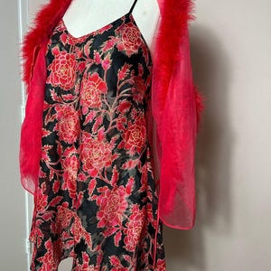 Könnte beinhalten: Ein schwarzes Slip-Kleid mit rotem Blumenmuster und Spaghettiträgern wird mit einer transparenten roten Federboa kombiniert. Das Kleid hat einen V-Ausschnitt und eine fließende Silhouette, die eine Vintage-Ästhetik erzeugt.
