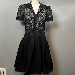 May include: A black dress with a lace bodice and short sleeves. The bodice has a V-neck and a floral lace design. The skirt is pleated and made of a smooth, shiny fabric. The dress is displayed on a mannequin.