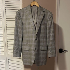May include: A grey and black plaid blazer featuring a notched lapel, two front buttons, and two flap pockets. The garment is constructed with long sleeves and a structured shoulder design, hanging on a wooden hanger with a solid grey interior lining.
