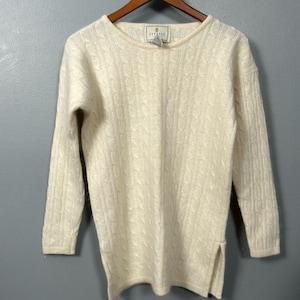 May include: Cream-coloured cable knit sweater with long sleeves. The sweater has a crew neck and a split hem on each side. The brand tag is visible at the neckline. The sweater is hanging on a wooden hanger.