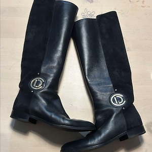 Puede incluir: Un par de botas negras hasta la rodilla. Las botas presentan un diseño de cuero y ante con un emblema metálico decorativo. Las botas tienen un tacón bajo y una silueta elegante, adecuadas para varios conjuntos. Aproximadamente 40 cm de altura.