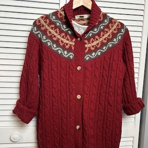 May include: A red cable-knit cardigan with a patterned yoke in beige, grey, and red. The button-down sweater has a shawl collar and three-quarter sleeves. The garment is hanging on a wooden hanger.