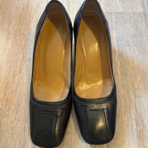 Può includere: Un paio di décolleté in pelle nera con design a punta quadrata. Le scarpe hanno un tacco medio e una finitura liscia e lucida. L'interno delle scarpe è di colore marrone chiaro. Le scarpe sono esposte su una superficie di legno chiaro.