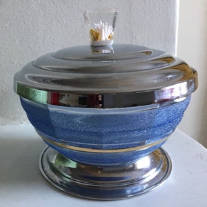 May include: A blue glass and chrome sugar bowl with a gold band around the middle. The lid has a white flower on top.