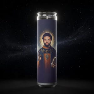 May include: A tall cylindrical prayer candle featuring a graphic illustration of a bearded man with a halo, wearing a dark blue and orange robe. The figure holds a brown American football in one hand and makes a hand gesture with the other.