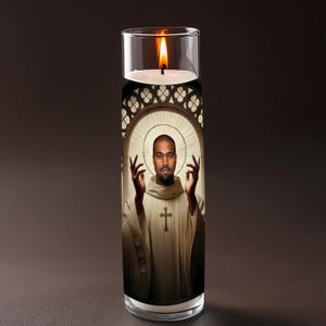 May include: A glass candle with a burning flame. The candle has an image of a man in a white robe with a cross, set against a stained-glass window design. The man is raising his hands.