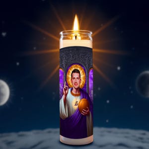May include: A lit prayer candle featuring a portrait of a man holding a basketball, with a halo and religious-style clothing. The candle has a purple and gold design with the letter "L" on the man's chest. The background is a night sky with stars and a moon.