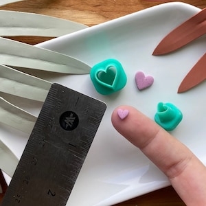 Puede incluir: Tres cortadores de galletas en forma de corazón turquesa y dos corazones de colores rosa sobre una superficie blanca. Una regla se encuentra en el fondo.