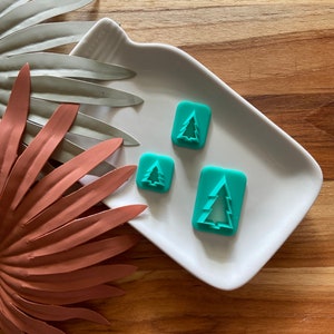 Può includere: Tre tagliapasta in plastica verde turchese a forma di alberi di Natale su un piatto bianco. Il piatto è su una superficie di legno.
