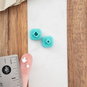 May include: Two turquoise blue plastic cookie cutters shaped like a bird, and two white plastic cookie cutters shaped like a bird, on a white surface with a wooden surface in the background.