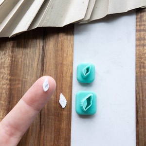 Puede incluir: Dos cortadores de galletas en forma de hoja de plástico turquesa sobre una superficie blanca. Un dedo sostiene un recorte de galleta en forma de hoja blanca.