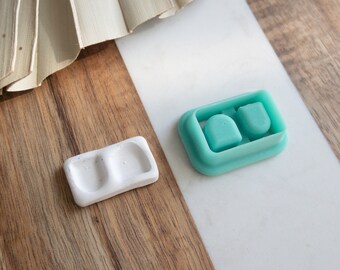 Puede incluir: Dos pequeños moldes de silicona blanca para hacer pendientes de resina, un molde está sobre una superficie de madera y el otro sobre una superficie blanca. Los moldes son ambos rectangulares con dos depresiones circulares.