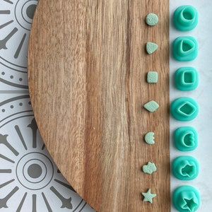 May include: A set of ten green plastic cookie cutters in various shapes, including a circle, a teardrop, a square, a leaf, a flower, a star, and a heart. The cutters are arranged in a row on a wooden chopping board.
