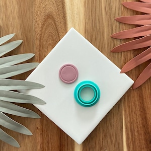 Könnte beinhalten: Zwei Fondant-Ausstecher auf einer weißen Oberfläche. Der Ausstecher links ist ein durchgehender Kreis und ist rosa. Der Ausstecher rechts ist ein Kreis mit drei Ringen und ist türkis.