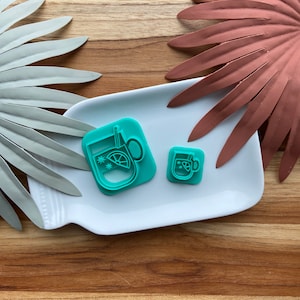 May include: Two teal blue cookie cutters shaped like mugs with a lemon slice and snowflakes inside. The cutters are on a white plate.
