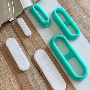 May include: Four teal plastic oval-shaped cookie cutters with a small handle on each side. The cutters are arranged on a wooden surface with three white oval-shaped pieces of clay.