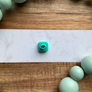 May include: A teal blue silicone mold shaped like a leaf with a small hole in the center. The mold is sitting on a white surface.