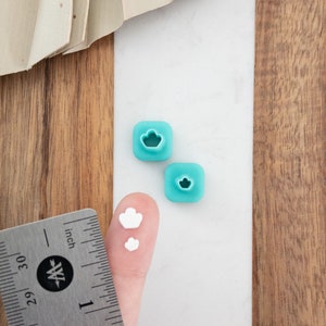 Peut inclure: Deux breloques carrées en argile polymère bleu turquoise avec un motif de nuage découpé et deux breloques en argile polymère blanche avec un motif de nuage. Une règle est en arrière-plan.