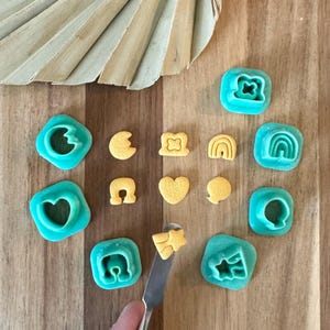 May include: A set of 10 teal blue plastic cookie cutters in various shapes, including a heart, a horseshoe, a rainbow, a star, and a crescent moon. The cutters are arranged on a wooden surface with yellow fondant cutouts.