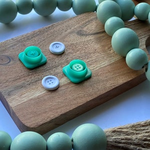 May include: Four green plastic buttons with white centers and four white plastic buttons with four holes each, on a wooden surface with a green beaded garland in the background.
