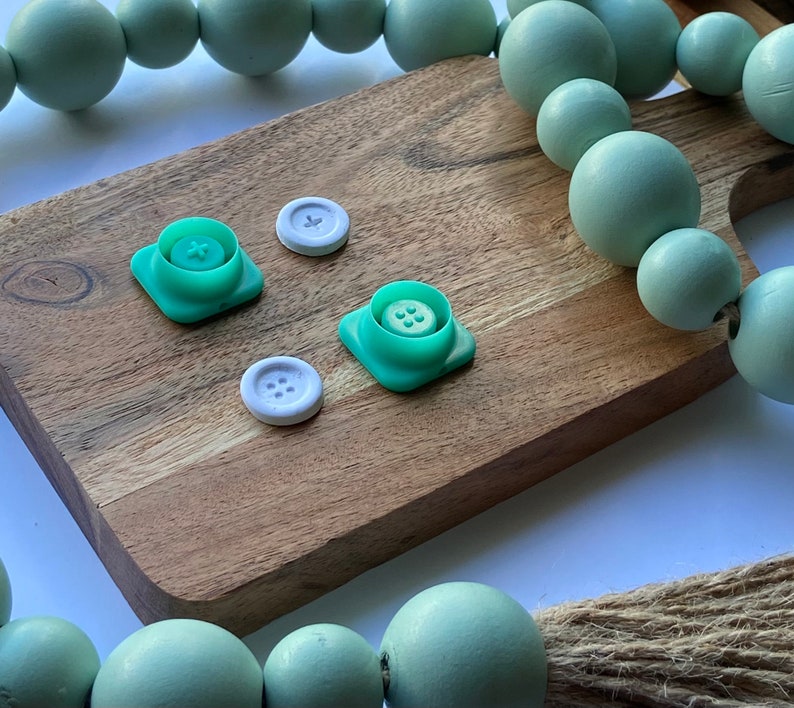 May include: Four green plastic buttons with white centers and four white plastic buttons with four holes each, on a wooden surface with a green beaded garland in the background.