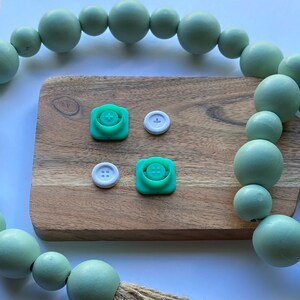 May include: Four green and white plastic buttons with different designs. The buttons are on a wooden surface with a green beaded garland in the background.
