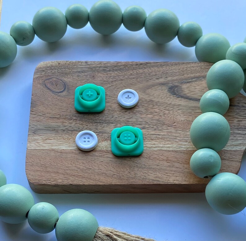 May include: Four green and white plastic buttons with different designs. The buttons are on a wooden surface with a green beaded garland in the background.