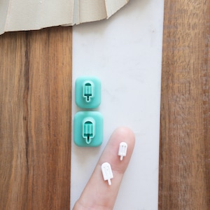 Może przedstawiać: Dwa turkusowe silikonowe foremki na lody na patyku z białym wzorem lodów na patyku. Palec trzyma dwa białe plastikowe lody na patyku.