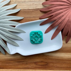 Puede incluir: Un cortador de galletas turquesa con un diseño geométrico del suroeste se encuentra sobre un plato rectangular blanco. El plato descansa sobre una superficie de madera, con acentos decorativos de hojas metálicas plateadas y cobrizas.