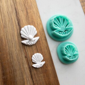 May include: Two white seashell-shaped embellishments and two teal seashell-shaped molds are displayed on a wooden surface. The embellishments have a pearl-like detail. The molds have a detailed seashell design.