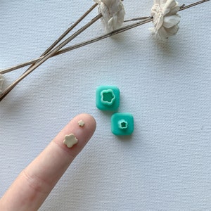 Puede incluir: Tres cortadores de galletas en forma de flor de plástico turquesa de diferentes tamaños. El cortador más pequeño está en un dedo, los otros dos están en una superficie blanca.