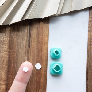 Puede incluir: Dos cortadores de galletas con forma de concha de plástico turquesa y dos conchas de fondant blanco sobre una superficie de madera.