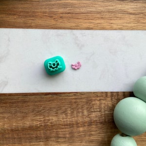 May include: Two small plastic cookie cutters, one shaped like a bow and the other shaped like a bow with a knot. The bow cutter is teal and the bow with a knot cutter is pink. The cutters are on a wooden surface.
