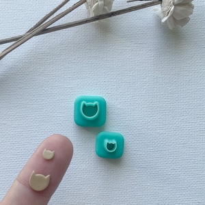 May include: Two turquoise blue plastic cat-shaped cookie cutters and a smaller beige cat-shaped cookie cutter.