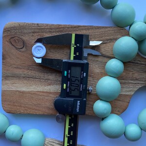 May include: A white button with a cross in the center is being measured with a digital caliper. The caliper reads 15.05 mm. The button is surrounded by a string of light green wooden beads.