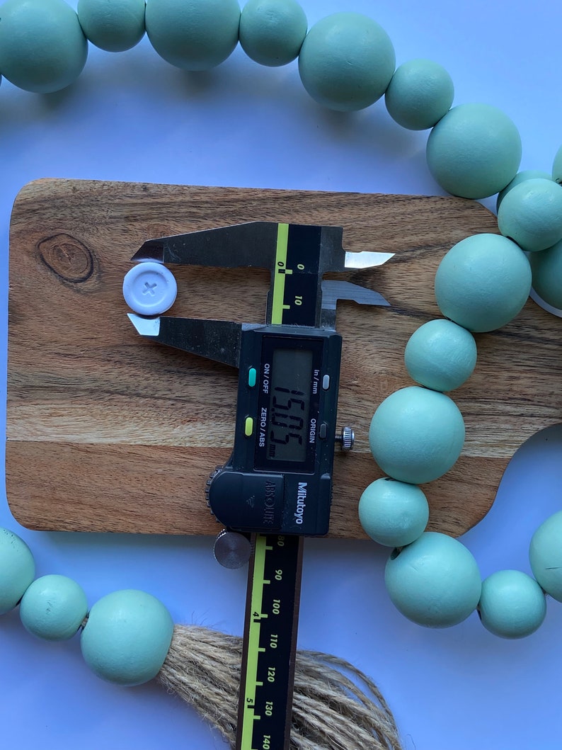 May include: A white button with a cross in the center is being measured with a digital caliper. The caliper reads 15.05 mm. The button is surrounded by a string of light green wooden beads.