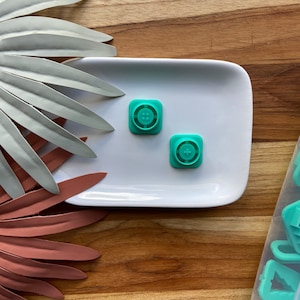 Peut inclure: Deux emportes-pièces en plastique bleu turquoise en forme de boutons, l'un avec quatre trous et l'autre avec un signe plus, sur une assiette blanche. L'assiette est sur une surface en bois.