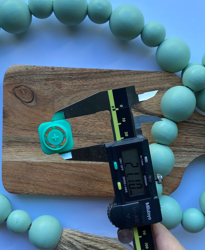 May include: A green plastic button with a plus sign in the center is being measured with a digital caliper. The caliper reads 21.10 mm.