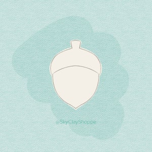 Puede incluir: Un contorno blanco de una bellota sobre un fondo azul claro con el texto "@SkyClayShoppe" en la parte inferior.