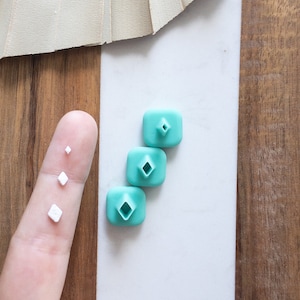 May include: Three turquoise blue square-shaped silicone bottle stoppers with a diamond-shaped hole in the center. The stoppers are arranged in a vertical stack, with the largest stopper at the bottom and the smallest stopper at the top.