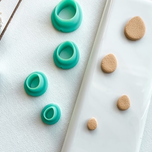 May include: Five teal-colored silicone molds for making polymer clay earrings. The molds are arranged in a row from largest to smallest. On the right side of the image, there are five tan-colored polymer clay circles, also arranged in a row from largest to smallest.