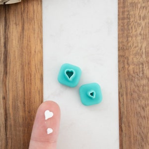 May include: Two turquoise blue heart-shaped cookie cutters with a small white heart on a fingertip.