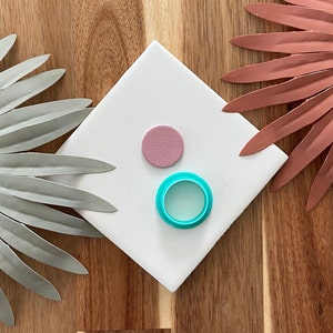 May include: A teal circle cutter and a small, round, pink object sit on a white square. The objects are surrounded by decorative, metallic, fan-shaped leaves in silver and copper colors, all on a wood-grain surface.