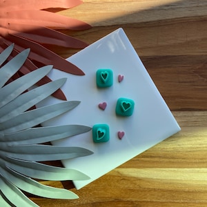 Puede incluir: Cuatro cortadores de galletas en forma de corazón de color azul verdoso sobre una superficie blanca con chispas de corazón rosa.