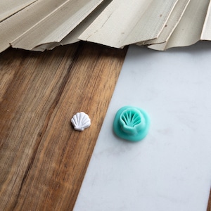 Può includere: Due stampini per fondant a forma di conchiglia, uno bianco e uno blu turchese. Gli stampini sono su una superficie di legno e marmo.