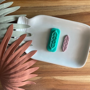 Puede incluir: Dos cortadores de galletas con forma de hoja, uno turquesa y otro morado, sobre un plato blanco. El cortador turquesa tiene un diseño geométrico y el cortador morado tiene un diseño texturizado.