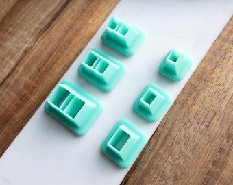 Puede incluir: Conjunto de seis herramientas de decoración de pasteles de plástico turquesa. Las herramientas son de diferentes tamaños y formas, con una base cuadrada y una abertura rectangular en la parte superior.