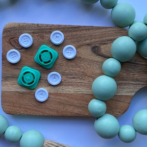May include: A wooden cutting board with white buttons and green plastic buttons. A light green beaded garland with a jute tassel hangs over the board.