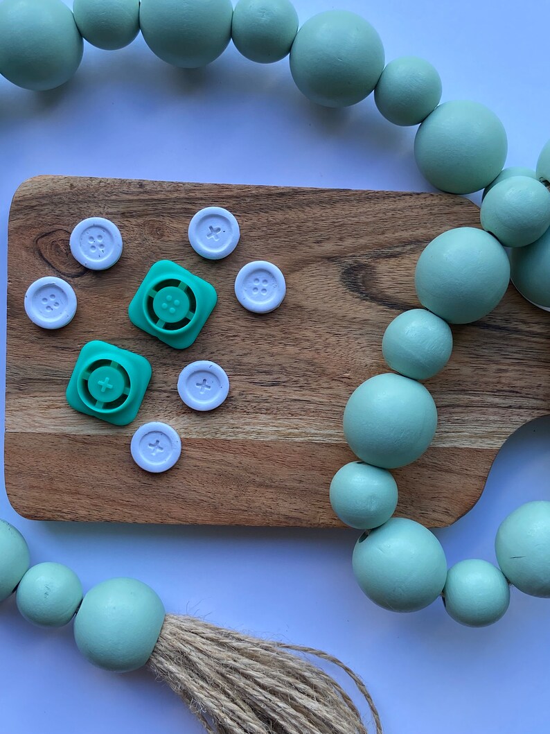 May include: A wooden cutting board with white buttons and green plastic buttons. A light green beaded garland with a jute tassel hangs over the board.